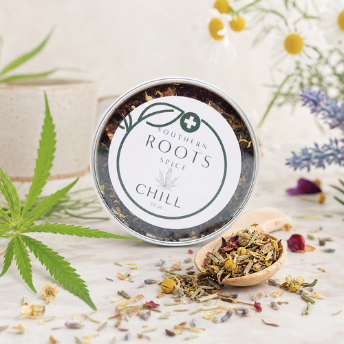 A round metal tin of Southern Roots Spice 'Chill' spice blend, shown with loose dried herbs in a small wooden spoon and scattered on a light surface, surrounded by decorative botanical props including a cannabis leaf, flowers, and a ceramic pot.