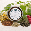 A round metal tin of Southern Roots Spice Comfort tea is styled on a light surface with loose tea ingredients and botanicals, including chamomile flowers, rosemary, green leaves, and raspberries.