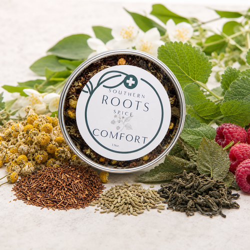 A round metal tin of Southern Roots Spice Comfort tea is styled on a light surface with loose tea ingredients and botanicals, including chamomile flowers, rosemary, green leaves, and raspberries.