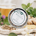 A round tin of Southern Roots Spice Company Detox Spice blend, styled on a stone surface with burdock root, mint, chamomile flowers, and a glass of tea in the background.