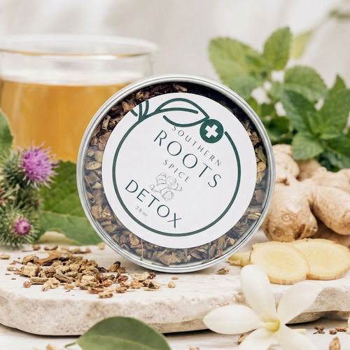 A round tin of Southern Roots Spice Company Detox Spice blend, styled on a stone surface with burdock root, mint, chamomile flowers, and a glass of tea in the background.