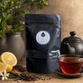 A black stand-up pouch of NY Tea Company's 'Forest Green' loose-leaf tea stands on a wooden table next to a glass cup of brewed tea, a teapot, lemon slices, loose tea leaves, and a small flower, with a blurred background.