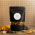 A black stand-up pouch of Southern Roots Spice Ginger Peach tea, displayed on a wooden surface with loose tea leaves, dried flowers, a metal tea infuser, and peach slices against a pegboard background.