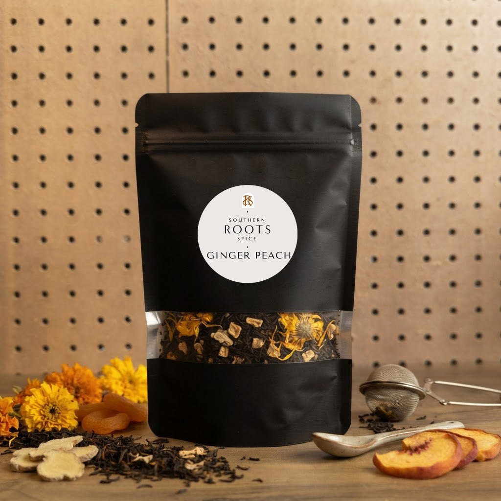 A black stand-up pouch of Southern Roots Spice Ginger Peach tea, displayed on a wooden surface with loose tea leaves, dried flowers, a metal tea infuser, and peach slices against a pegboard background.