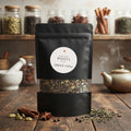 A black stand-up pouch of Southern Roots Spice Green Chai tea with a clear window showing the loose leaf blend, set on a rustic wooden table with spices and a teapot in the background.