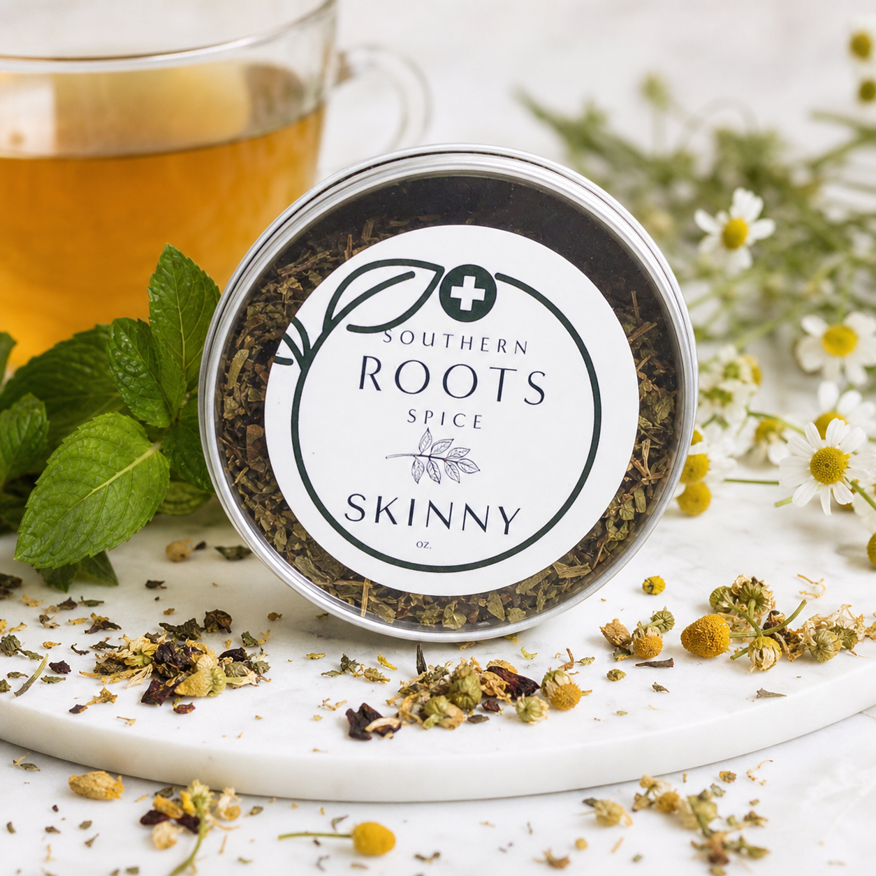 A round metal tin of Southern Roots Spice Skinny spice blend sits on a white marble board with loose herbs and flowers around it. A glass mug of amber tea and green mint leaves are blurred in the background.