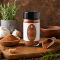 A bottle of Southern Roots Spice Southern Rib Rub seasoning stands on a wooden cutting board with bowls of the spice, garlic, rosemary, and peppercorns around it.